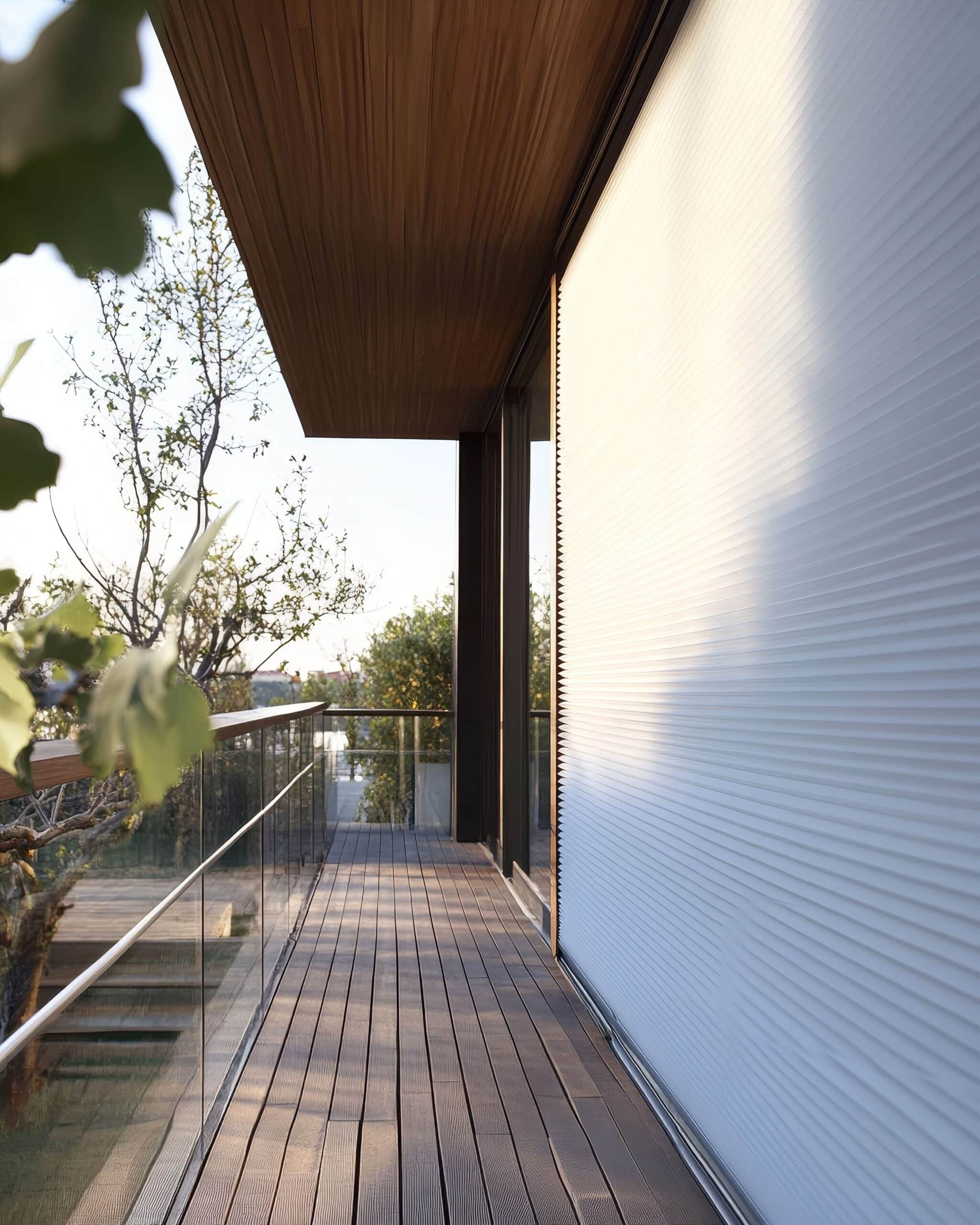 Terrasse en bois design, balustrade en verre, arbres luxuriants et store plissé blanc baigné de lumière naturelle.