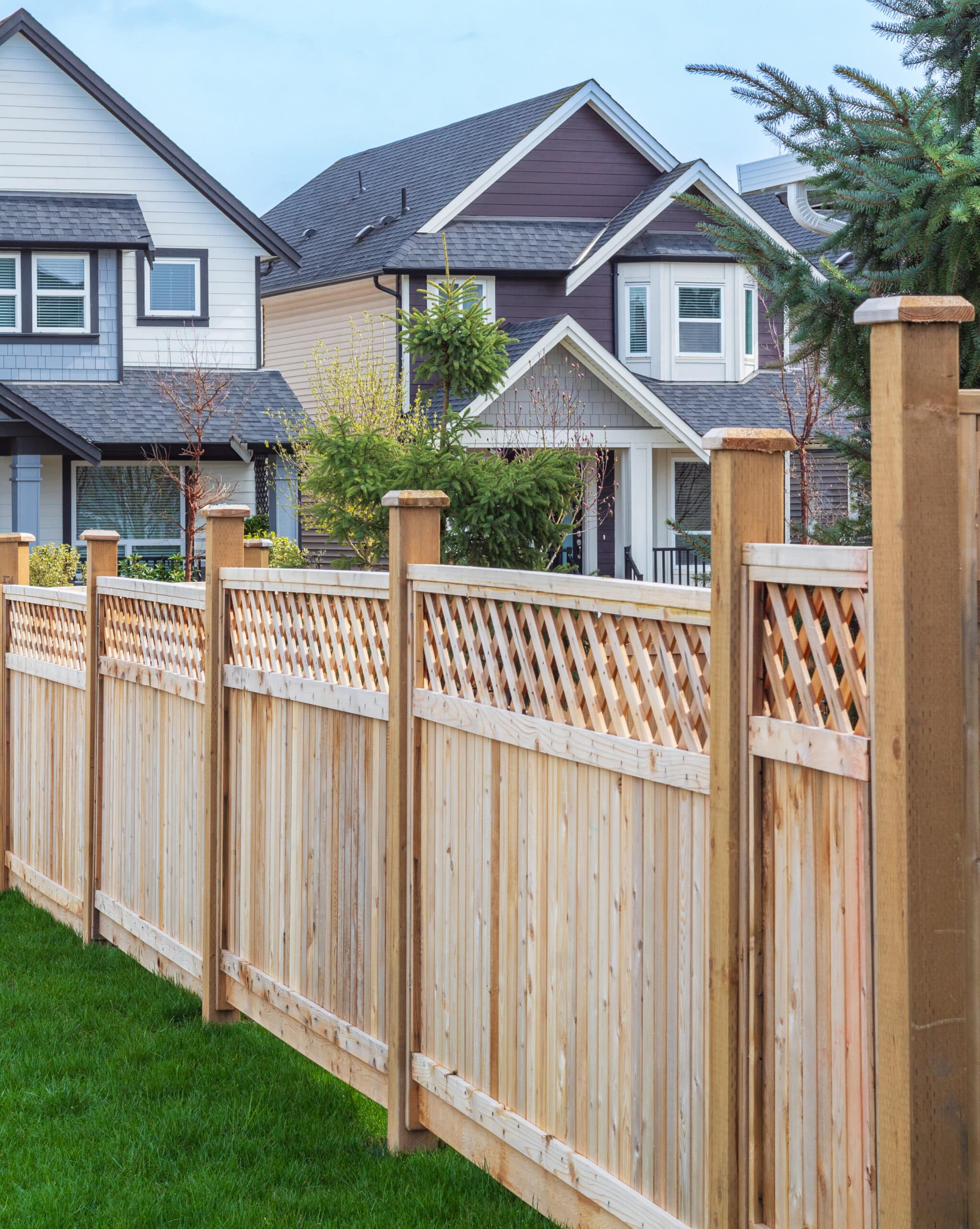 Clôture en bois neuve avec treillis et pelouse verte au premier plan. Maisons résidentielles modernes et arbres en arrière-plan sous ciel clair.