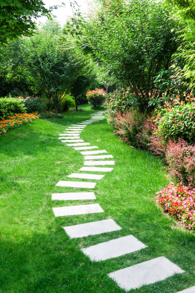 Chemin de dalles en pierre serpente dans un jardin luxuriant. Pelouse verte, arbres et buissons variés, fleurs oranges et rouges vif.