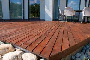 Grande terrasse moderne en bois avec mobilier de jardin devant une maison blanche et vitrée. Reflets d'arbres et ciel bleu.