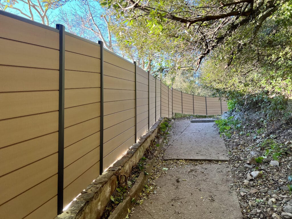 Longue clôture moderne en bois clair avec poteaux sombres, serpentant le long d'un sentier extérieur bordé d'arbres et de verdure sous un ciel bleu.