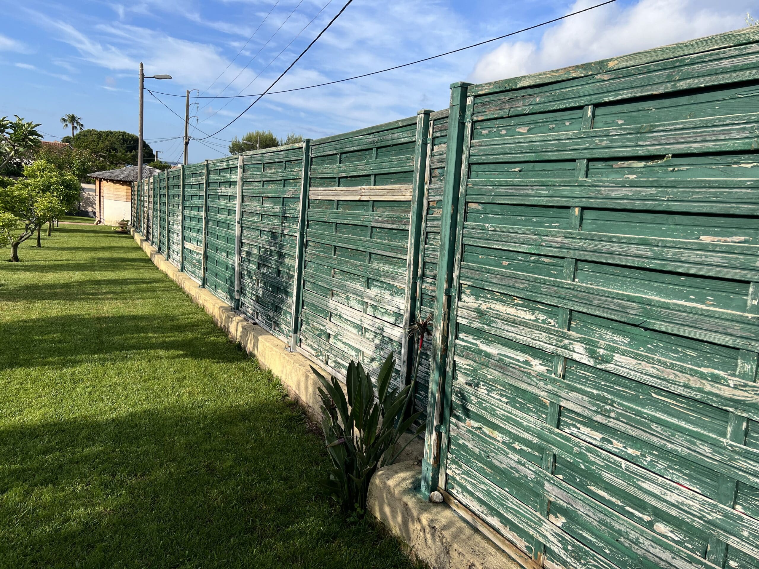 Longue clôture de jardin en bois vert écaillé longeant une pelouse luxuriante. Au-delà, ciel bleu, arbres, et poteaux électriques.