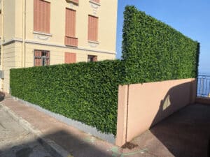 Façade jaune, volets corail, haie végétale artificielle dense. Mur rose avec chaîne rouillée sur trottoir. Mer Méditerranée et ciel bleu.