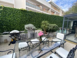 Patio avec table à manger en verre, chaises noires, coussins blancs, roses. Barbecue, plantes, haie verte, vélos turquoise, bâtiment en arrière-plan sous ciel bleu.