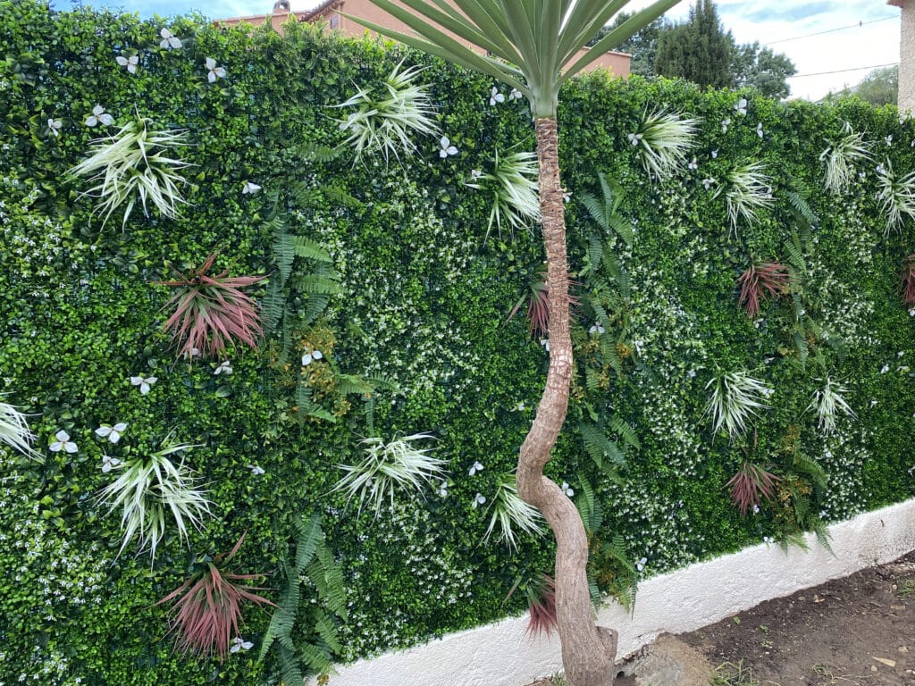 Mur végétal artificiel vert luxuriant avec fougères, plantes spikées et fleurs blanches. Tronc de Yucca courbé en premier plan.