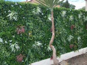 Mur végétal artificiel vert luxuriant avec fougères, plantes spikées et fleurs blanches. Tronc de Yucca courbé en premier plan.