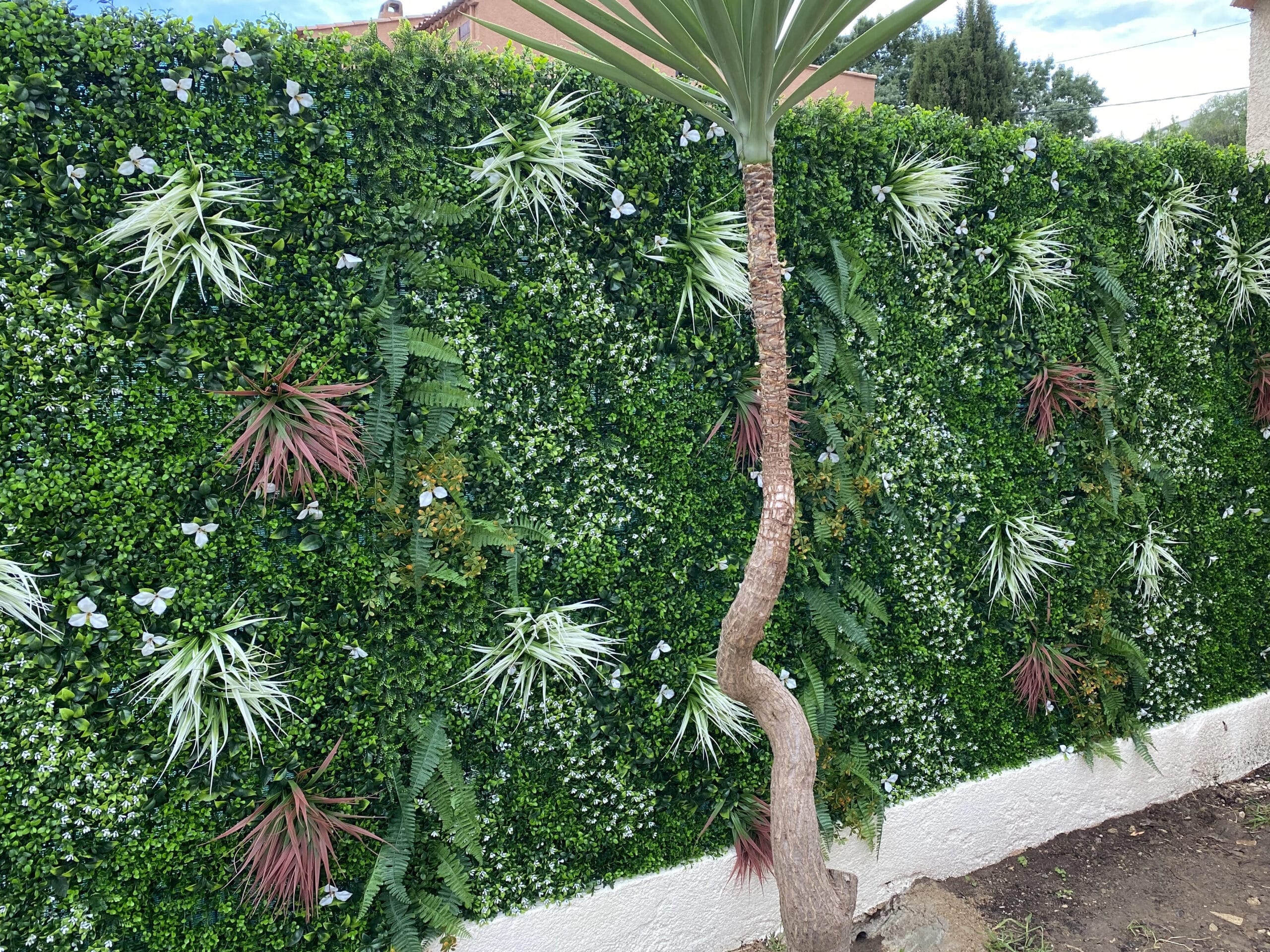 Mur végétal artificiel vert luxuriant avec fougères, plantes spikées et fleurs blanches. Tronc de Yucca courbé en premier plan.