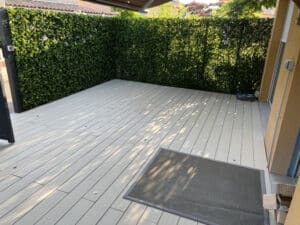 Terrasse moderne en composite gris clair, haie artificielle verte avec guirlande lumineuse. Ombre d'arbres sur le sol, tapis gris extérieur.