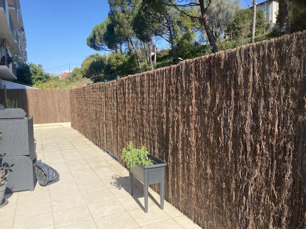 Terrasse carrelée ensoleillée avec salon de jardin gris, grande clôture en brande, plantes vertes en pot et ciel bleu.